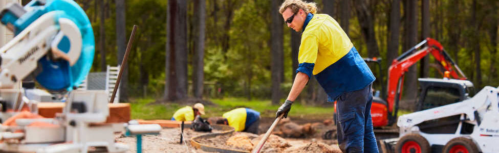 Worker installing metal lawn edging on a residential landscape project, surrounded by tools, timber, and earthmoving equipment in a wooded area.